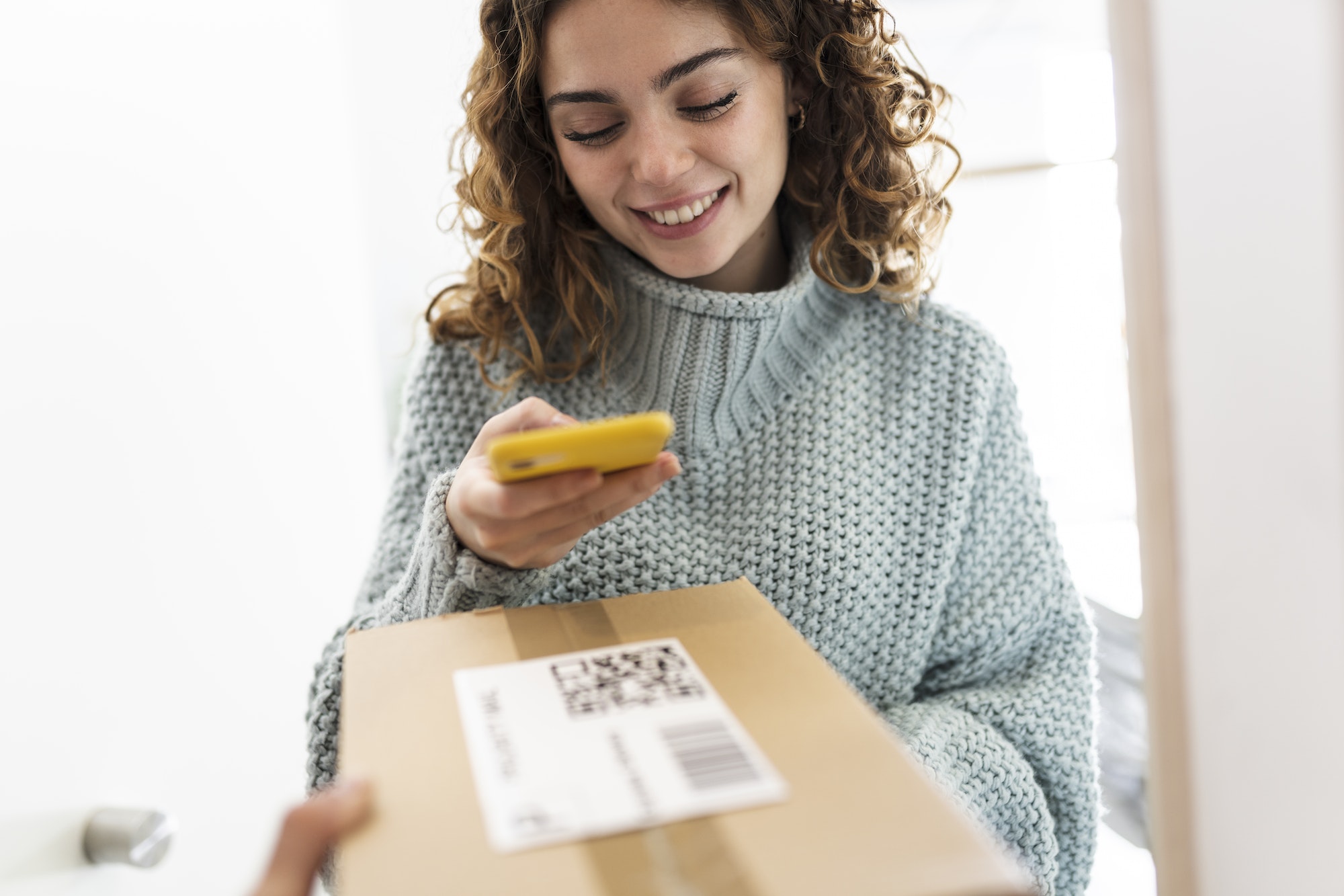 Woman with curly hair scanning a QR code with her smartphone to receive a home delivery package.